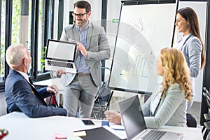 Handsome young businessman holding laptop and giving presentation to his business colleagues in boardroom office.