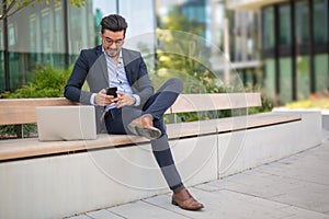 Handsome young businessman with laptop texting sms on his smartphone