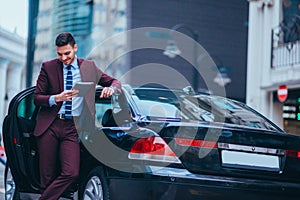 Handsome young businessman leaning on his limo and smiling while looking at his phone