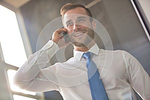 Handsome young businessman smiling while talking on smart phone while standing in new office