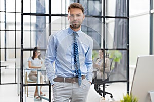 Handsome young businessman standing in modern office with collegues on the background.