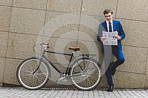 handsome young businessman in stylish suit with bicycle reading newspaper leaning