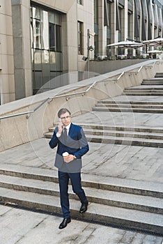 handsome young businessman in stylish suit with coffee to go talking by phone while going