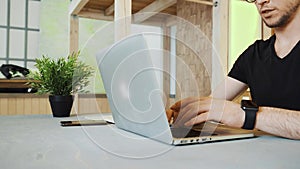 Handsome young businessman working in loft office. A focused young man with glasses turns on his laptop and starts typing.