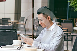 Handsome young caucasian man sitting at cafe using mobile phone and having coffee. Asian male with cup of coffee and reading text