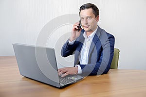 Handsome young concentrated attractive man using cellphone while working with laptop in the office