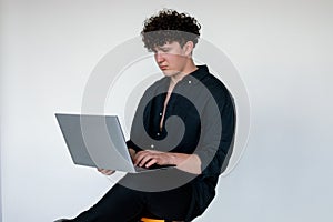 Handsome young curly guy in total black look sitting on chair and playing computer games on modern laptop, gray studio