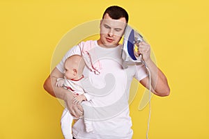 Handsome young dad looking after newborn baby, needs ironing, holds iron near ear like mobile phone, imagines he talks to somebody