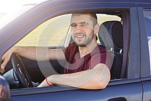 Handsome young dark haired male with stubble dressesd maroon casual t shirt, poses in black car, looks at camera trough window,