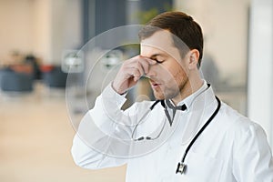 Handsome young doctor in white coat is touching his temples while standing in hospital hall.
