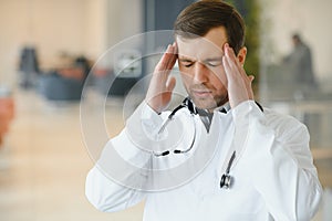 Handsome young doctor in white coat is touching his temples while standing in hospital hall.