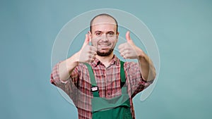 a handsome young farmer with a big smile and a thumb up is captured wearing overalls in a studio shot.