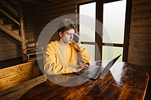 Handsome young freelancer man working in a wooden country house on a laptop and calling by phone. Freelancer working on a laptop