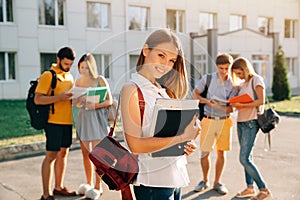 Handsome young girl with red velvet backpack holding books and smiling while standing against university with her friends in the