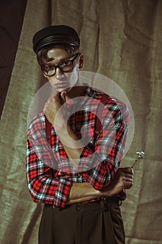 Handsome young guy in a cap and checkered shirt is holding a wild flower in his hand