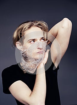 A handsome young guy with long blonde hair and sad blue eyes holds a bubble wrap in front of his face on a gray background