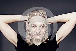 A handsome young guy with long blonde hair and sad blue eyes holds a bubble wrap in front of his face on a gray background