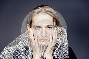 A handsome young guy with long blonde hair and sad blue eyes holds a bubble wrap in front of his face