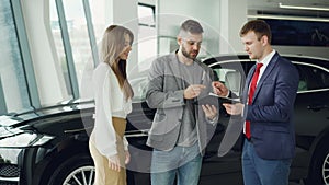 Handsome young guy is signing sale and purchase agreement in car dealership, getting key fob from salesman and giving it