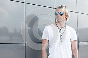 Handsome young hipster blond man in blue round sunglasses with stylish hairstyle in a trendy white t-shirt with amulets is