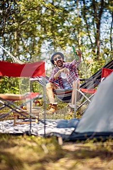 Handsome young hipster male cheering with beer and sitting in hammock. Picnic in forest. Holiday, leisure, fun, lifestyle concept