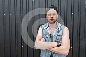 Handsome young hipster man in a fashionable blue denim vest in a t-shirt with a beard in a black stylish cap poses