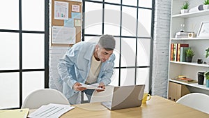 Handsome young hispanic man engrossed in reading business documents on laptop at office, hardworking and focused on success