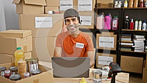 Handsome young hispanic man selflessly volunteering, working on laptop with headphones at charity center, donations box nearby