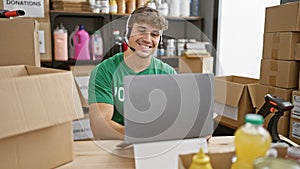 Handsome young hispanic man volunteering at a community charity center, cheerfully smiling while sitting at a table, using laptop