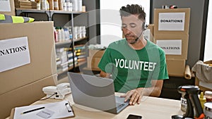 Handsome young hispanic man volunteering at community charity center, sitting at table, working online on laptop with headphones,