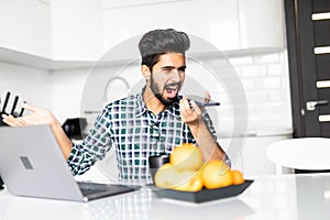 Handsome young indian man angry talking on phone while working at table in kitchen