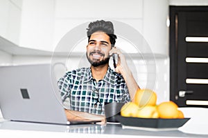 Handsome young indian man talking on phone while working at table in kitchen