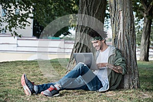 Handsome young international student sitting on grass in public park, typing text message on keyboard of modern laptop computer,