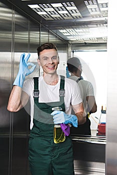 handsome young janitor holding spray bottle with detergent and rag smiling at camera and showing ok sign