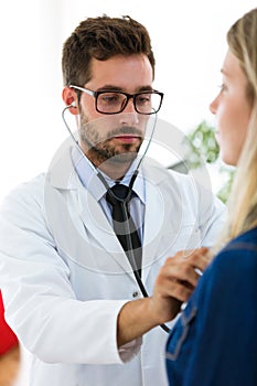 Handsome young male doctor checking beautiful young woman patient heartbeat using stethoscope in medical office.
