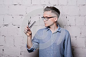 Handsome young male hairdresser in glasses, posing with scissors and comb, on gray brick wall background, horizontal