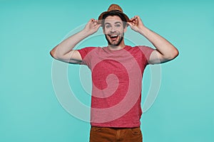 Handsome young man adjusting his fedora hat and smiling while standing against blue background