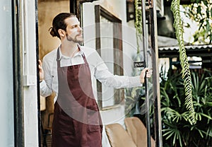 Handsome young man in apron working waitress in cafe opening entrance door and inviting customers to his shop. Cheerful owner of