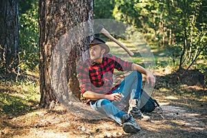 Handsome young man with axe near forest. Lumberjack sitting in the forest.