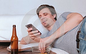 Handsome young man bachelor eating chips and drinking beer and browsing his mobile phone.