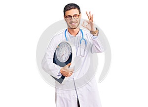 Handsome young man with bear as nutritionist doctor holding weighing machine doing ok sign with fingers, smiling friendly