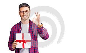 Handsome young man with bear holding gift doing ok sign with fingers, smiling friendly gesturing excellent symbol