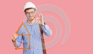 Handsome young man with bear wearing architect hardhat holding build project doing ok sign with fingers, smiling friendly
