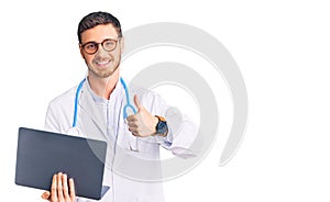 Handsome young man with bear wearing doctor uniform working using computer laptop smiling happy and positive, thumb up doing