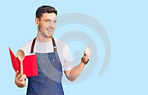Handsome young man with bear wearing professional baker apron reading cooking recipe book pointing thumb up to the side smiling