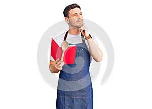 Handsome young man with bear wearing professional baker apron reading cooking recipe book serious face thinking about question