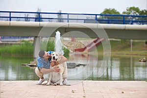 Handsome young man with beard and his Labrador retriever dog, posing for photos by a lake in the park. The man shows his affection