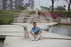 Handsome young man with beard and his Labrador retriever dog, posing for photos by a lake in the park. The man shows his affection