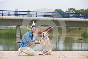 Handsome young man with beard and his Labrador retriever dog, posing for photos by a lake in the park. The man shows his affection