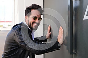 Handsome young man with beard, sculpted body and sunglasses is leaning with both hands on the grey door of his garage looking at